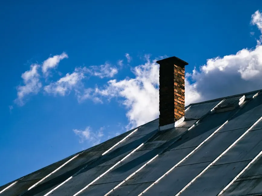 A scenic view of a metal roof with a brick chimney under a clear blue sky adorned with fluffy white clouds.